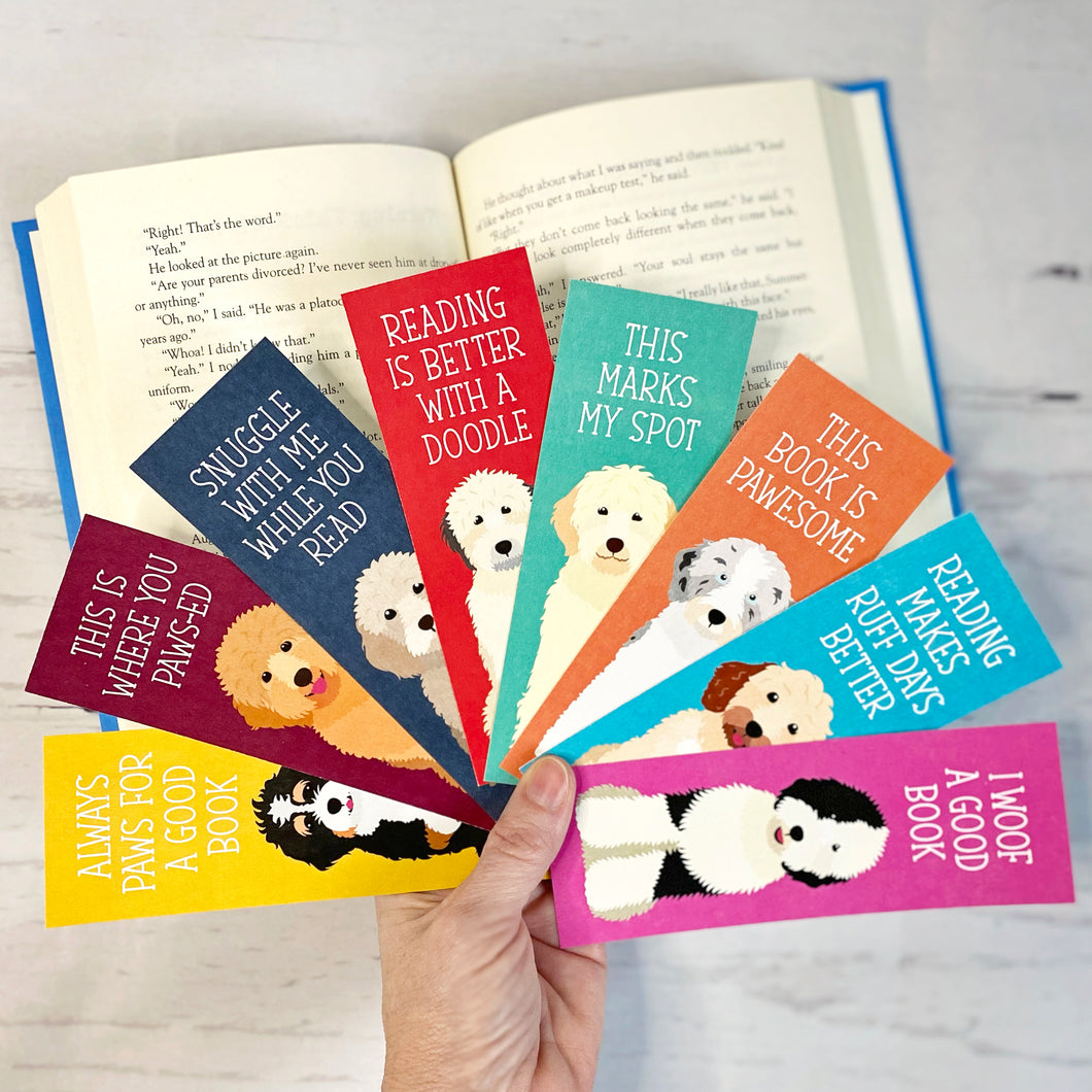 8 colorful book marks with adorable illustrations of different doodle dogs on each one with cute doggy sayings. A hand is holding all of the bookmarks in a fan over an open book
