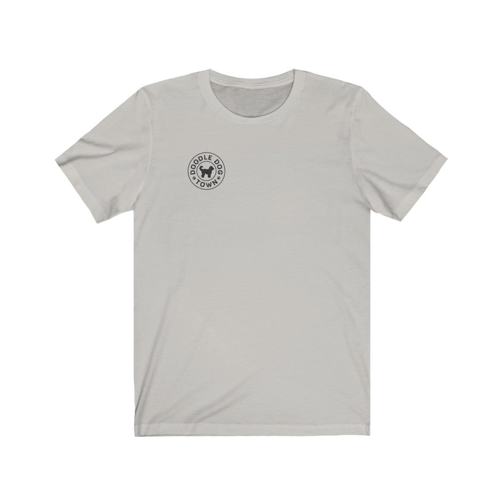 A small black logo of Doodle Dog Town which consists of a circle with the words inside following the shape of the circle with a black silhouette of a doodle dog in the middle on a silver t-shirt