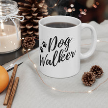 Load image into Gallery viewer, white ceramic mug, handle on right side that reads &quot;Dog Walker&quot; with two paws all in black. Mug sitting on table cloth with three pine cones and a string of fairy lights and cinnomon sticks in the foreground. Holiday theme 
